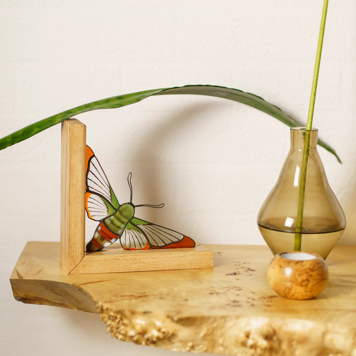 A decorative stained glass hummingbird moth decor piece displayed on a rustic wooden shelf next to a small glass vase with a green stem.