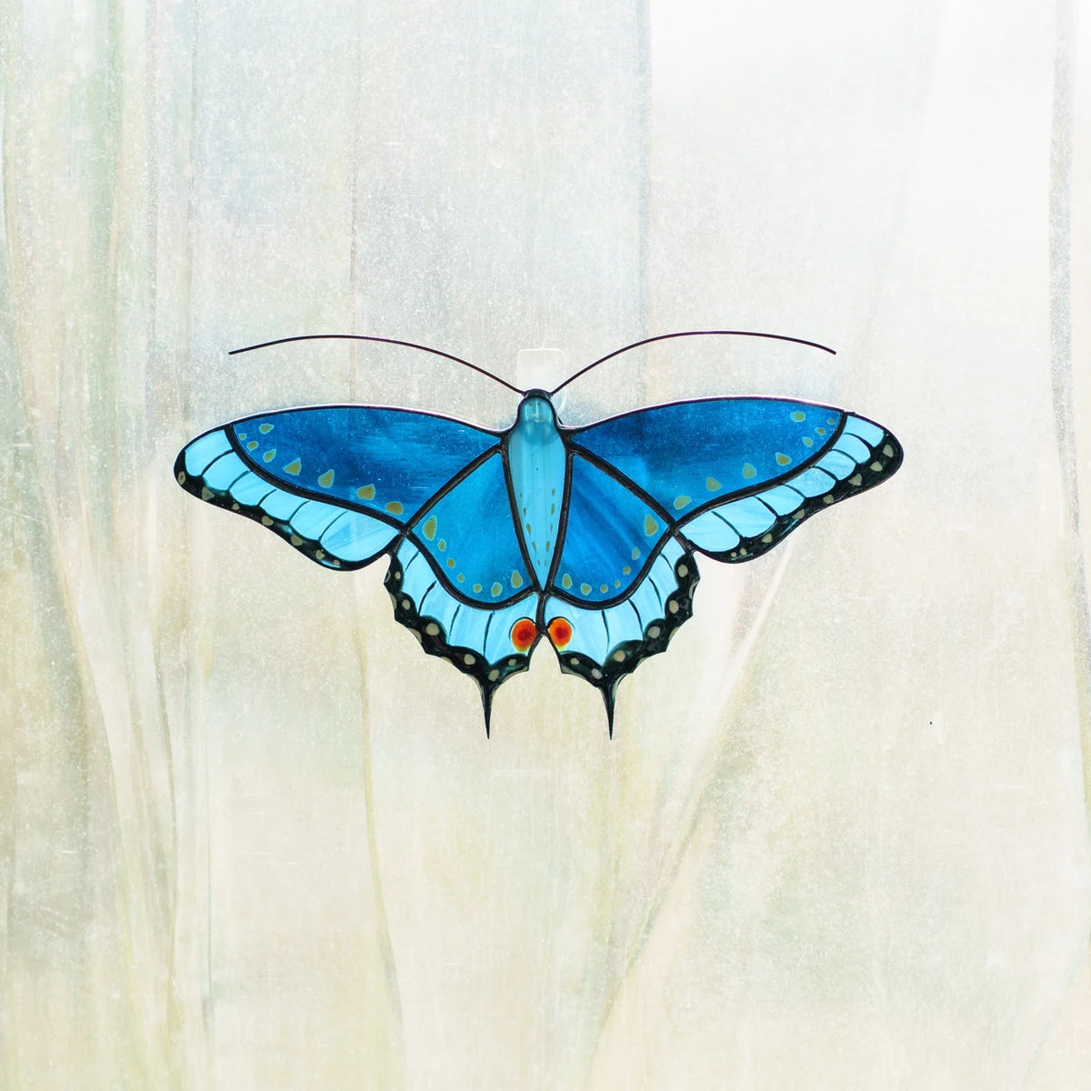 Blue swallowtail stained glass suncatcher attached to a window with a clear adhesive hook, daylight behind it