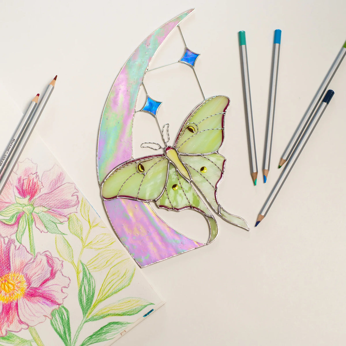 Luna moth and moon stained glass on a white table beside a peony drawing and colored pencils