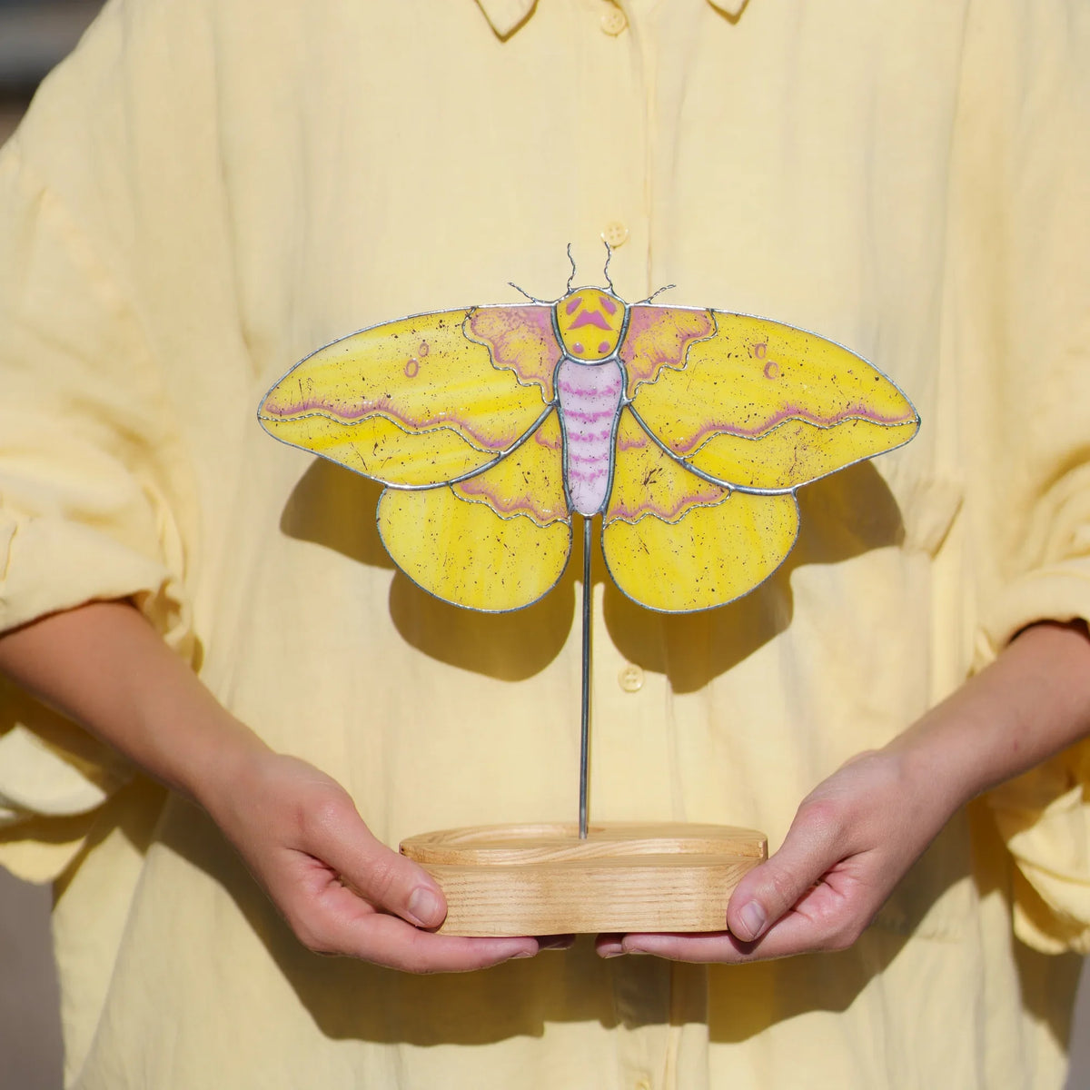 Person holding the imperial moth stained glass suncatcher by its engraved wooden base
