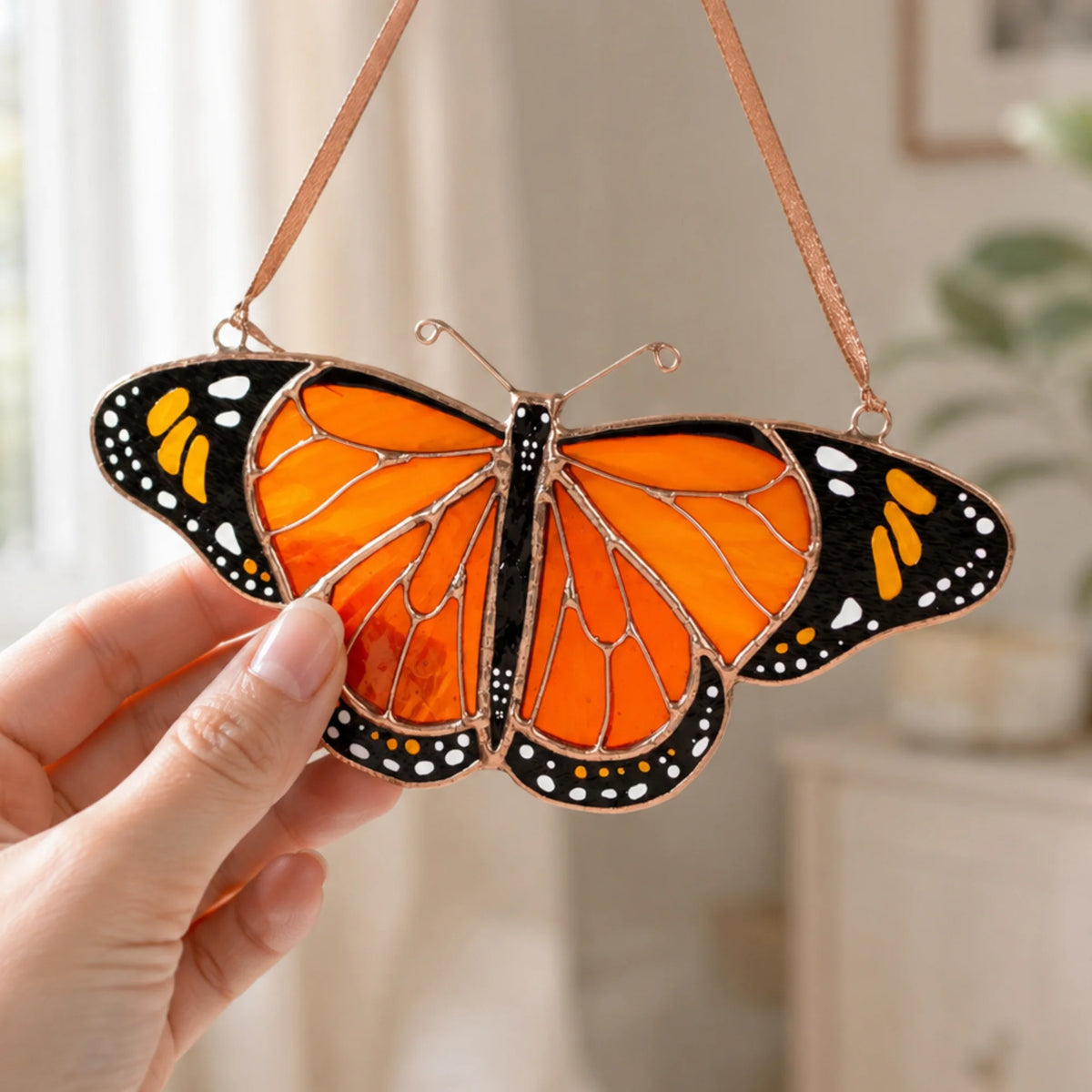 A hand holds a stained-glass butterfly ornament with bright orange wings, black borders, white spots, and copper-toned metal lines and hanging straps, against a softly blurred indoor background.