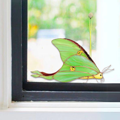 A green moth-shaped stained glass window ornament with yellow eyes and antennae, attached to a window pane.
