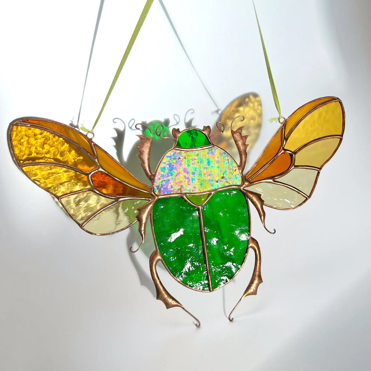 A stained glass scarab beetle ornament with a vibrant green body, iridescent multicolor back, and translucent yellow, orange, and cream wings, suspended by thin green strings against a white background.