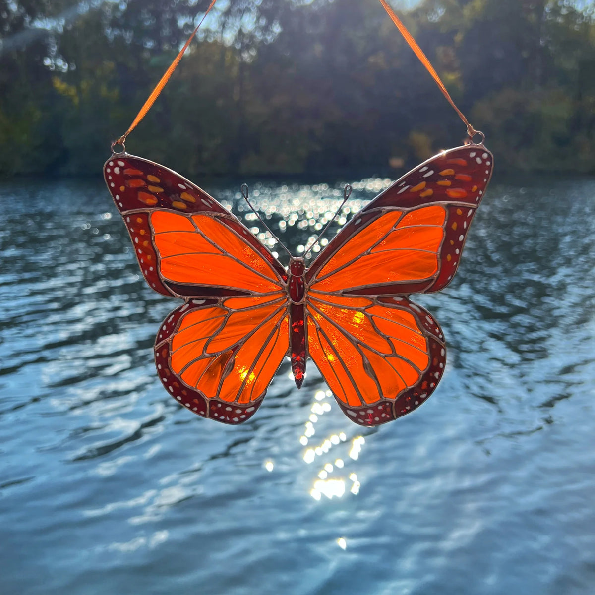An orange butterfly stained glass suncatcher with a hanging ribbon, photographed outdoors against a blurred lake and trees background.
