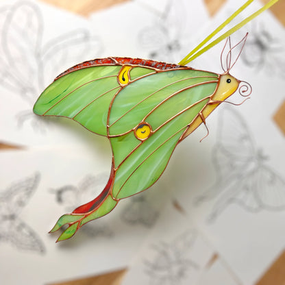 A stained glass luna moth ornament with green wings, yellow eyes, and copper wire details, shown against a blurred background of sketch drawings.