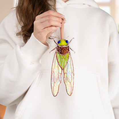 Green stained glass cicada hanging from a ribbon, rainbow sheen across its iridescent stained glass wings and amber textured glass thorax