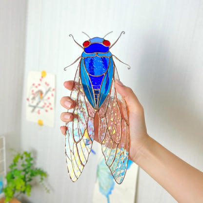 Hand holds a stained glass cicada ornament with a bright blue body, red eyes, and patterned wings in a softly lit indoor setting.