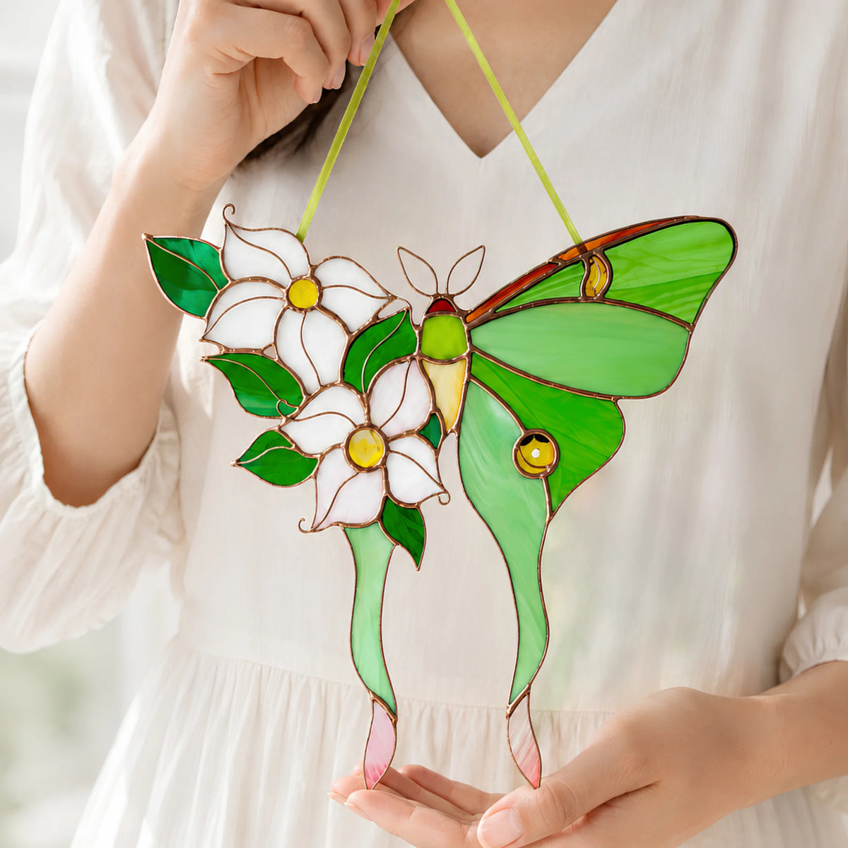 A stained glass hanging ornament shaped like a green moth with white flowers and green leaves, held by a person wearing a white blouse.