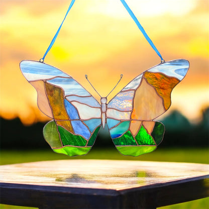 A stained glass butterfly ornament with a Yosemite landscape design, suspended by blue strings above a wooden table against a warm sunset sky.