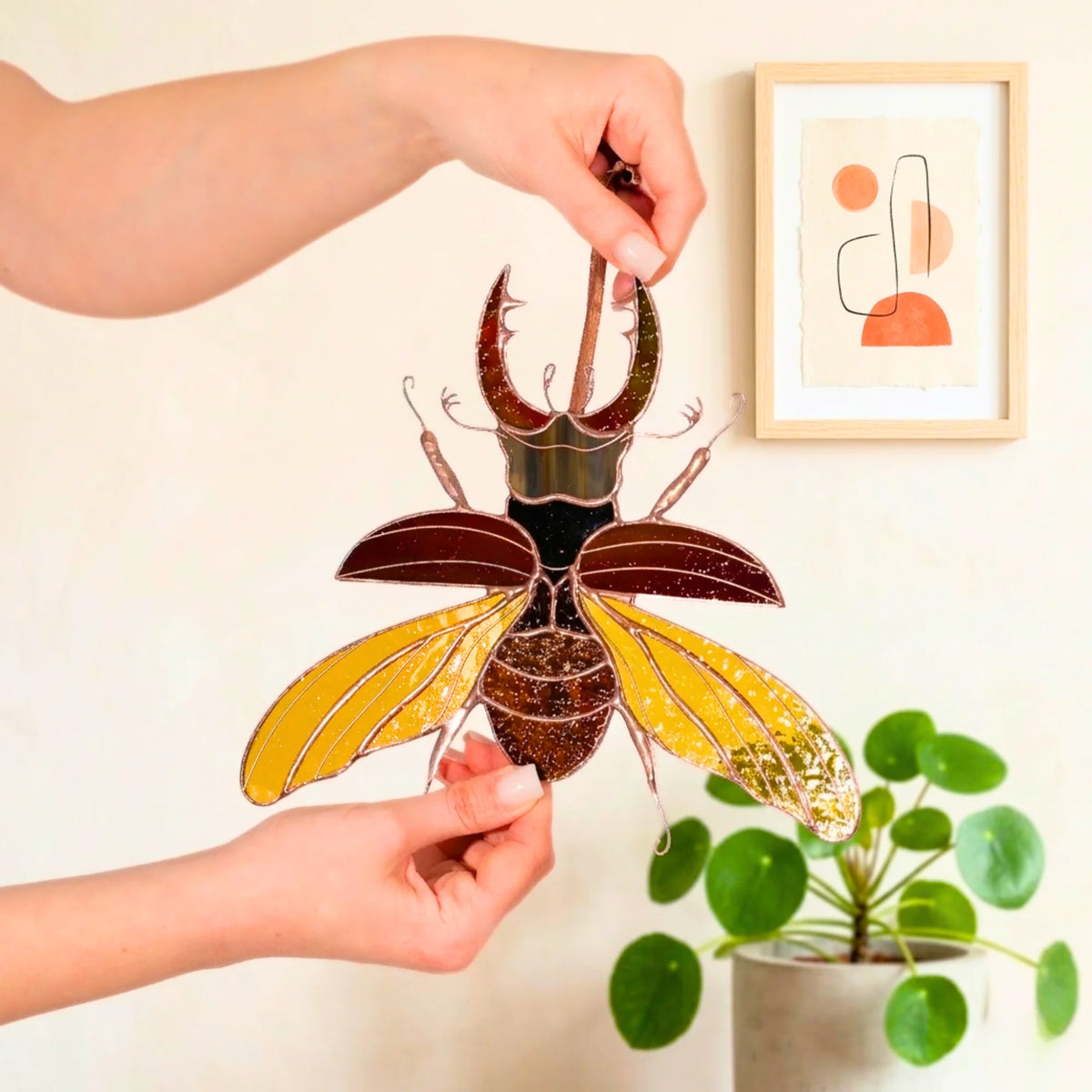 A person holds a decorative stag beetle stained glass ornament with metallic gold and dark brown sections, shown against a light wall with a framed abstract print and a potted plant in the background.