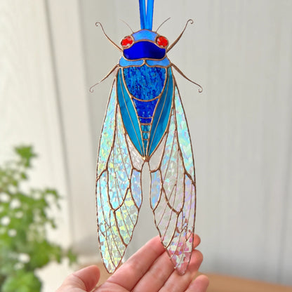 A stained glass cicada ornament being held in a person's hand, featuring iridescent wings, a bright blue body, red eyes, and a blue hanging ribbon against a softly blurred indoor background.