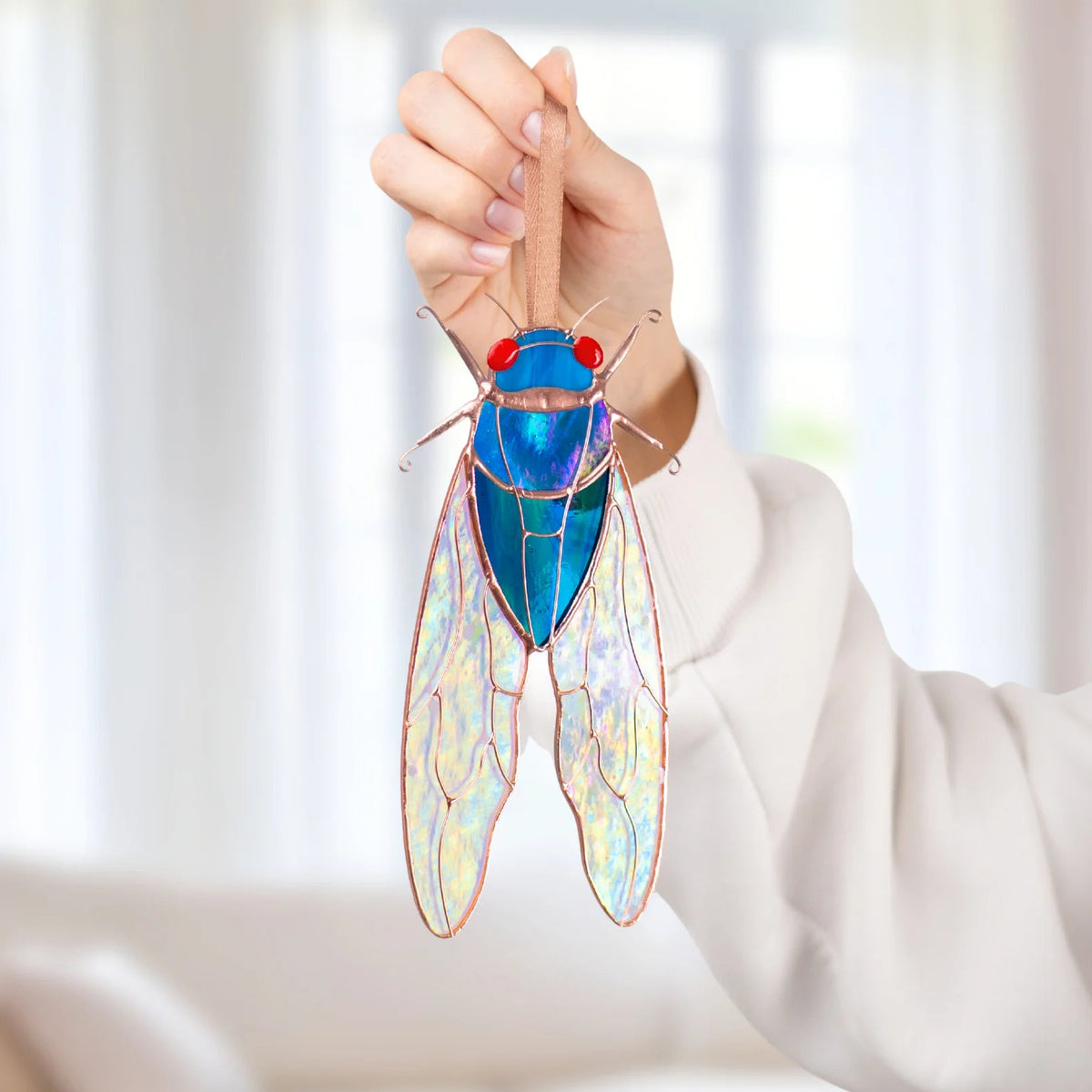 Hand holding a blue stained glass cicada window hanging by its ribbon, pastel light shining through the iridescent stained glass wings