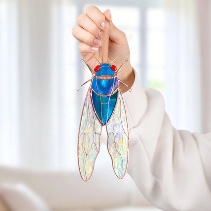 Hand holding a blue stained glass cicada window hanging by its ribbon, pastel light shining through the iridescent stained glass wings