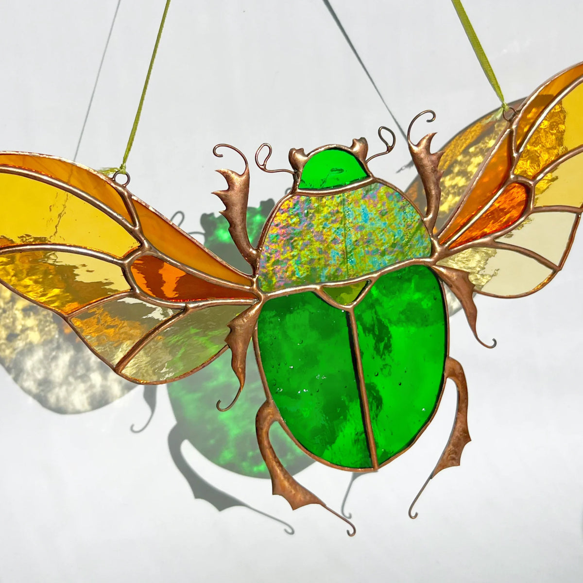 A stained glass scarab beetle suncatcher with a bright green body and translucent wings in yellow, orange, and cream, suspended by thin green strings against a white background.