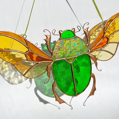 A stained glass scarab beetle suncatcher with a bright green body and translucent wings in yellow, orange, and cream, suspended by thin green strings against a white background.