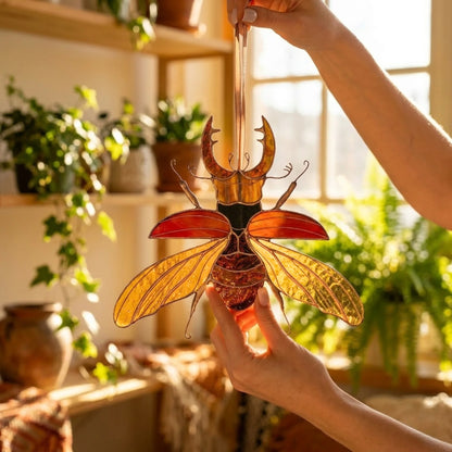 A handcrafted stained glass stag beetle suncatcher with amber, orange, and red glass panels, held by two hands in a sunlit room with plants in the background.
