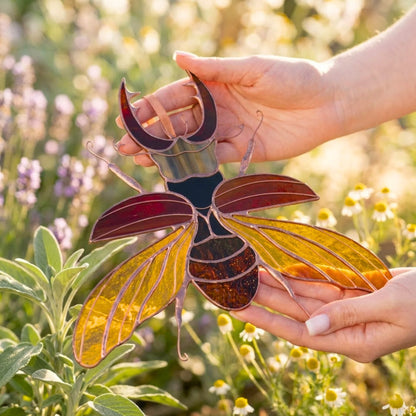 A stained glass stag beetle suncatcher with amber, gold, and dark red glass panels is held in two hands against a soft-focus outdoor garden background.