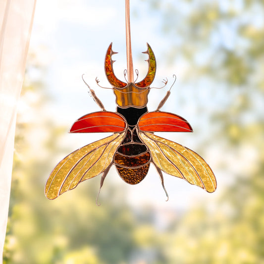 A stained glass beetle ornament hanging in front of a bright window. The beetle features metallic gold wings, a black and gold body, and reddish-orange wings, with a hanging cord at the top.