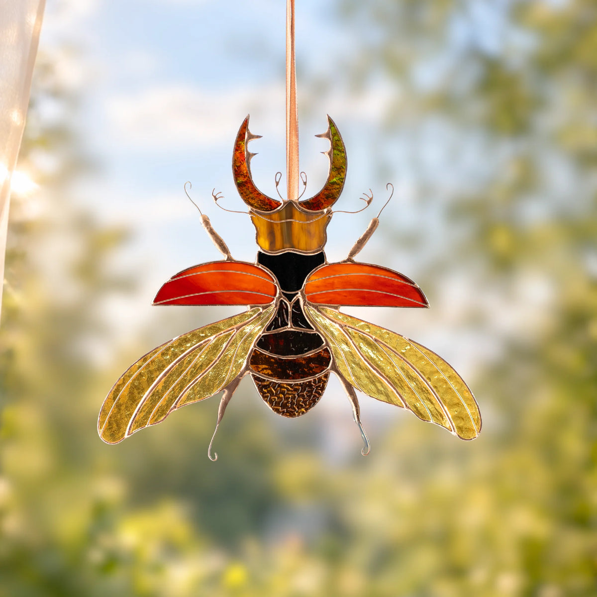 A stained glass beetle suncatcher hanging in front of a bright, sunlit window. The beetle features metallic gold and bronze wings, a black and gold body, and reddish-orange elytra, set against a blurred green and blue outdoor background.