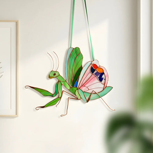 Stained glass hanging wall decor piece shaped like a praying mantis with a butterfly wing, made from green glass panels and gold-toned metal frames, hanging on a white wall next to a framed botanical print.