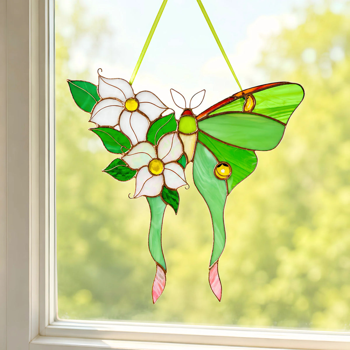 A stained glass hanging ornament a green luna moth butterfly with white flowers and green leaves, suspended by yellow-green strings in front of a window with a blurred outdoor background.
