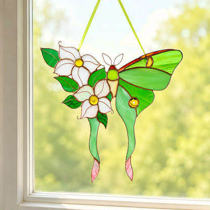 A stained glass hanging ornament a green luna moth butterfly with white flowers and green leaves, suspended by yellow-green strings in front of a window with a blurred outdoor background.