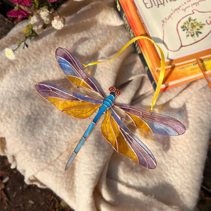 A stained glass dragonfly ornament with a blue body, purple and gold wings, and a yellow hanging ribbon, displayed on a beige fabric surface next to an open book.