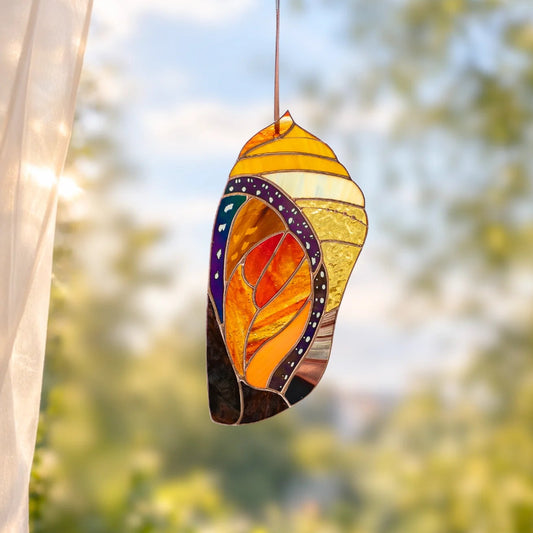 A stained glass suncatcher shaped like a monarch chrysalis in front of a window. The leaf design features yellow, orange, red, purple, and black sections with a hanging string at the top.