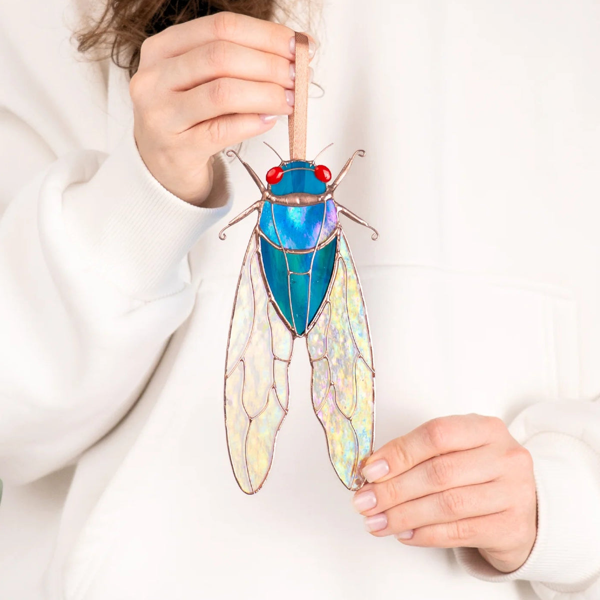 Woman in a white hoodie holding a blue stained glass cicada suncatcher with both hands to show its length