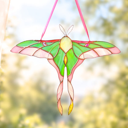 A stained glass hanging suncatcher shaped like a large moth or butterfly, with green, pink, cream, and yellow panels outlined in metallic gold-toned lines, suspended by pink ribbons against a soft blurred outdoor background.