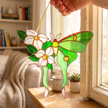 A stained glass window ornament featuring green luna moth holding white flowers, held by a hand in front of a sunlit window.