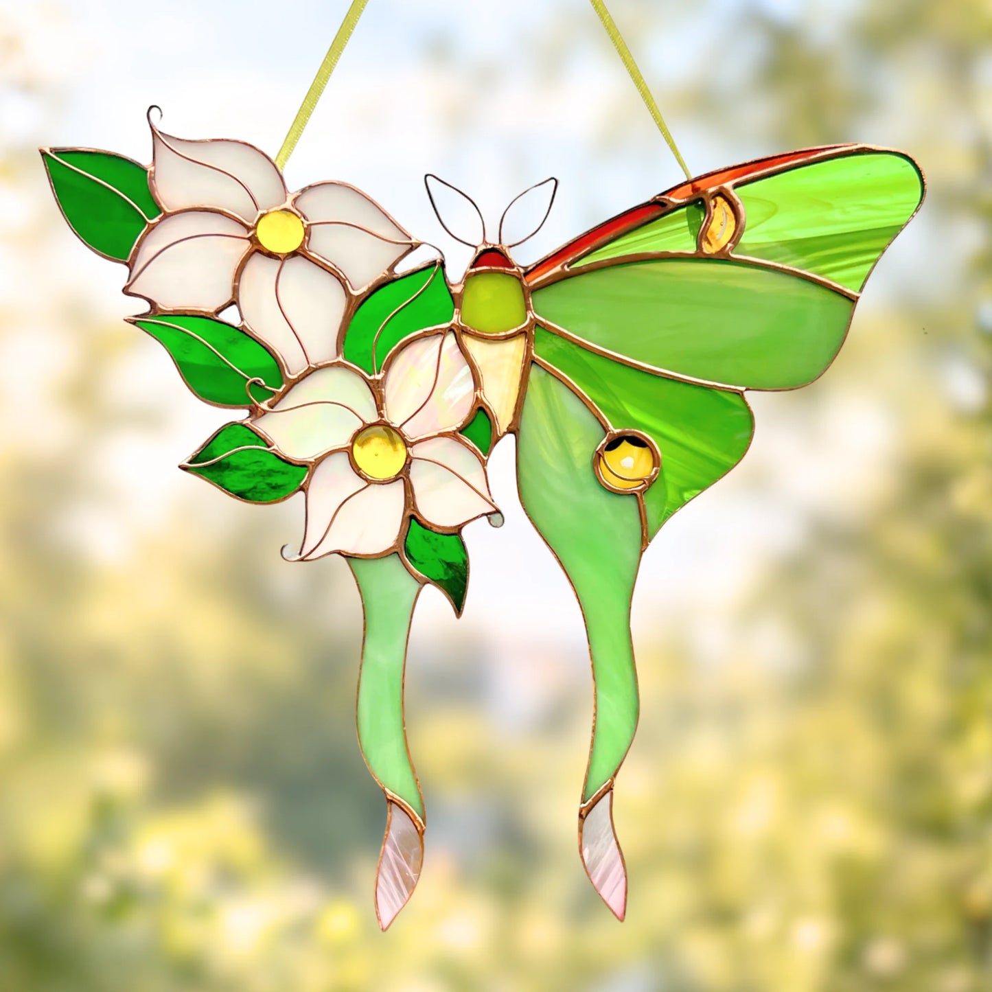 A stained glass hanging ornament featuring a green luna moth with two white flowers and green leaves, suspended by thin cords against a soft, blurred outdoor background.
