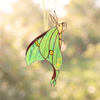 A stained glass suncatcher shaped like a luna moth, featuring vibrant green wings with yellow and brown details, hanging in front of a soft-focus, sunlit outdoor background.