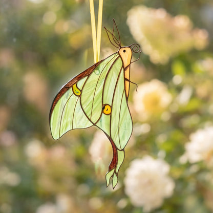 A stained glass luna moth ornament hanging from a thin yellow string against a soft, blurred garden background of green foliage and white flowers.