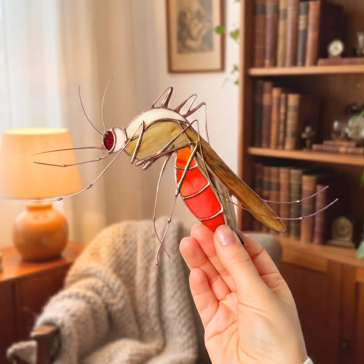A hand holds a stained glass mosquito window ornament in a cozy, warmly lit room with a bookshelf and lamp in the background.
