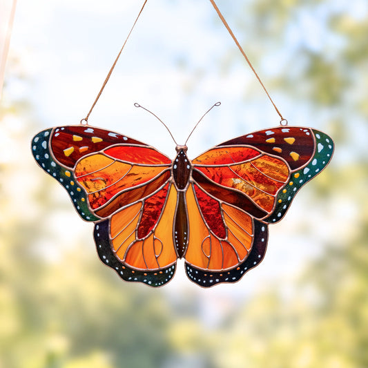A stained glass butterfly suncatcher with vibrant orange, red, and brown wings, accented with black, green, and white details, hanging by thin metal wires against a soft, blurred green and yellow outdoor background.