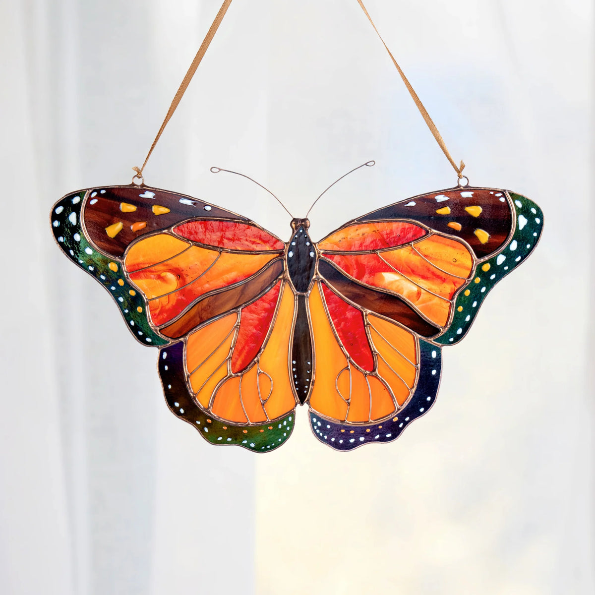 A stained glass butterfly ornament, hanging by a thin string against a soft white curtain background. The monarch butterfly features vibrant orange and yellow wings with dark brown borders, green dotted edges, and dark blue sections on the lower wings.