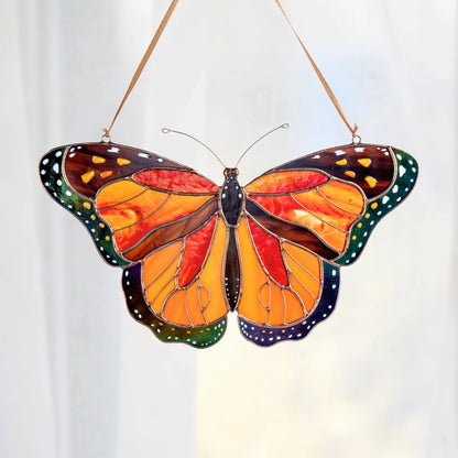 A stained glass butterfly ornament, hanging by a thin string against a soft white curtain background. The monarch butterfly features vibrant orange and yellow wings with dark brown borders, green dotted edges, and dark blue sections on the lower wings.