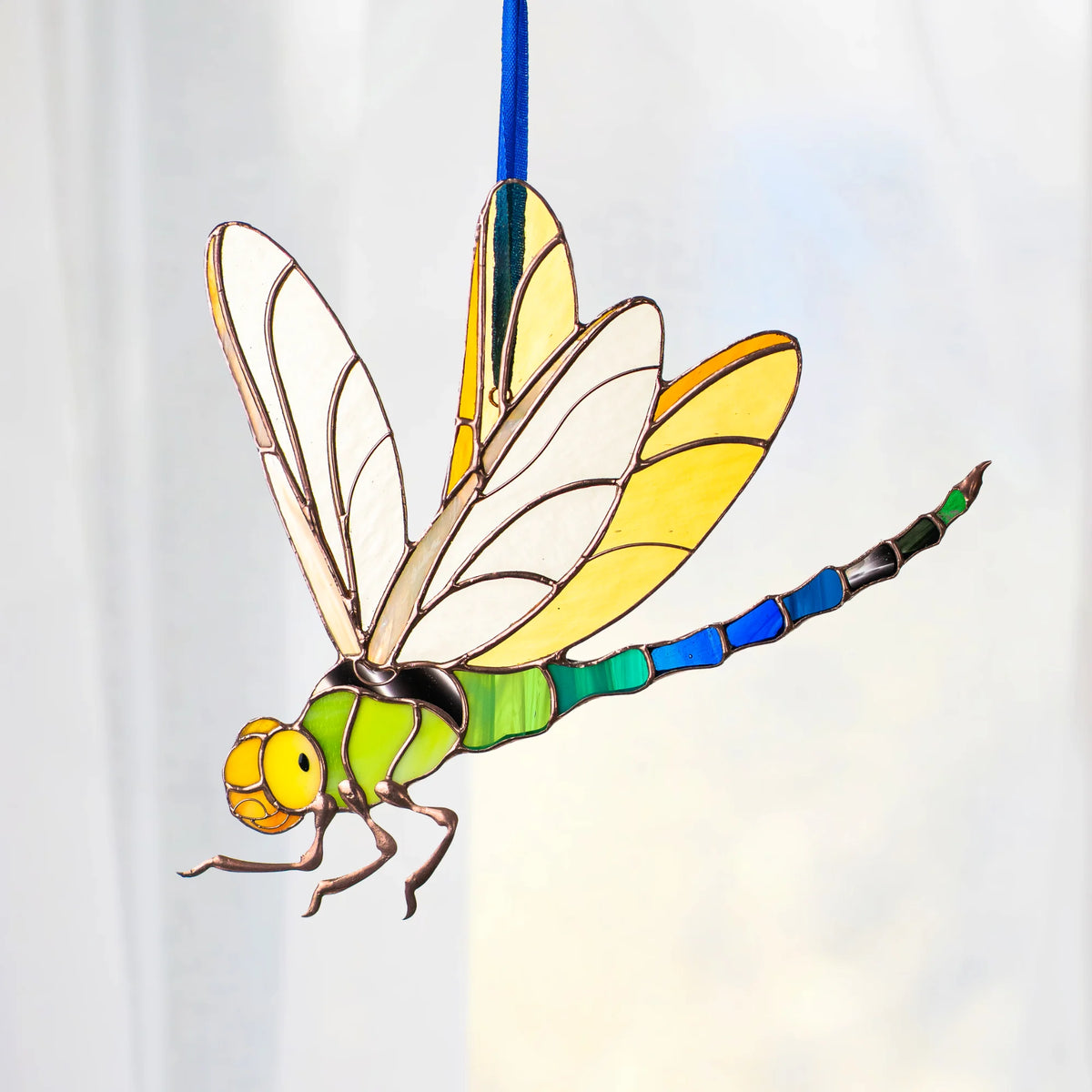 A stained-glass style dragonfly ornament or decoration with a green body, yellow and white wings, blue segmented tail, and gold-toned outlines, shown against a soft white background.
