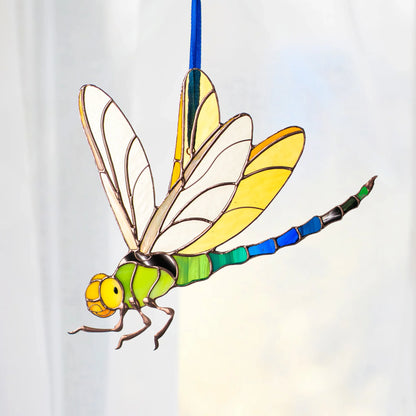 A stained-glass style dragonfly ornament or decoration with a green body, yellow and white wings, blue segmented tail, and gold-toned outlines, shown against a soft white background.