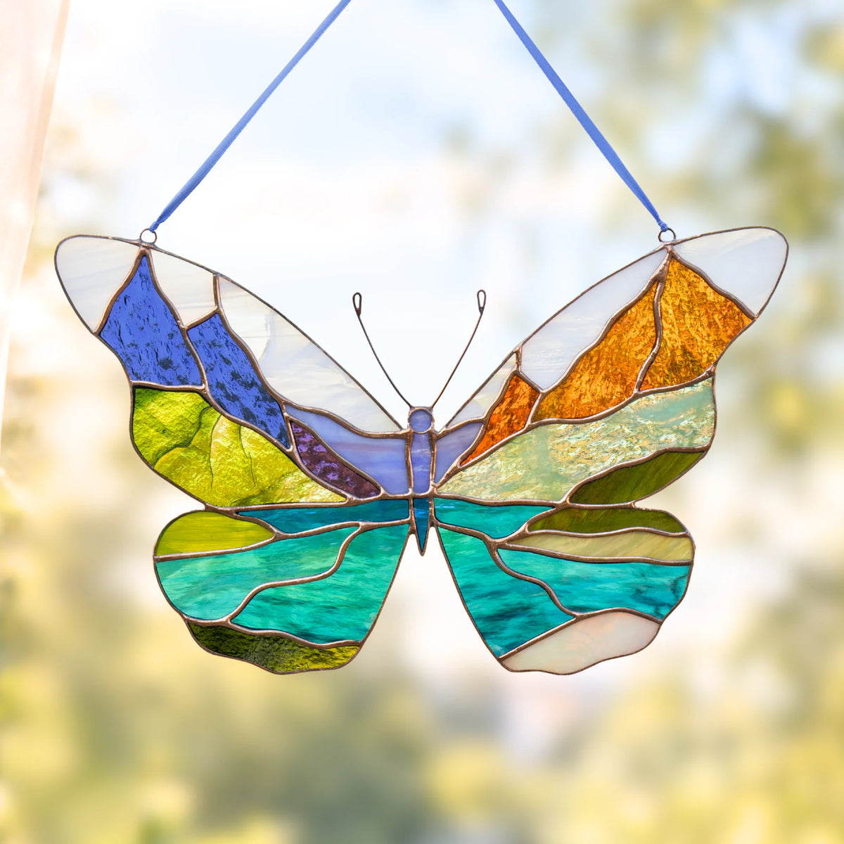 A stained glass butterfly suncatcher hanging by blue strings, featuring Glacier made of blue, green, yellow, orange, and purple glass against a soft, blurred outdoor background.