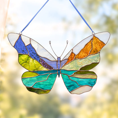 A stained glass butterfly suncatcher hanging by blue strings, featuring Glacier made of blue, green, yellow, orange, and purple glass against a soft, blurred outdoor background.
