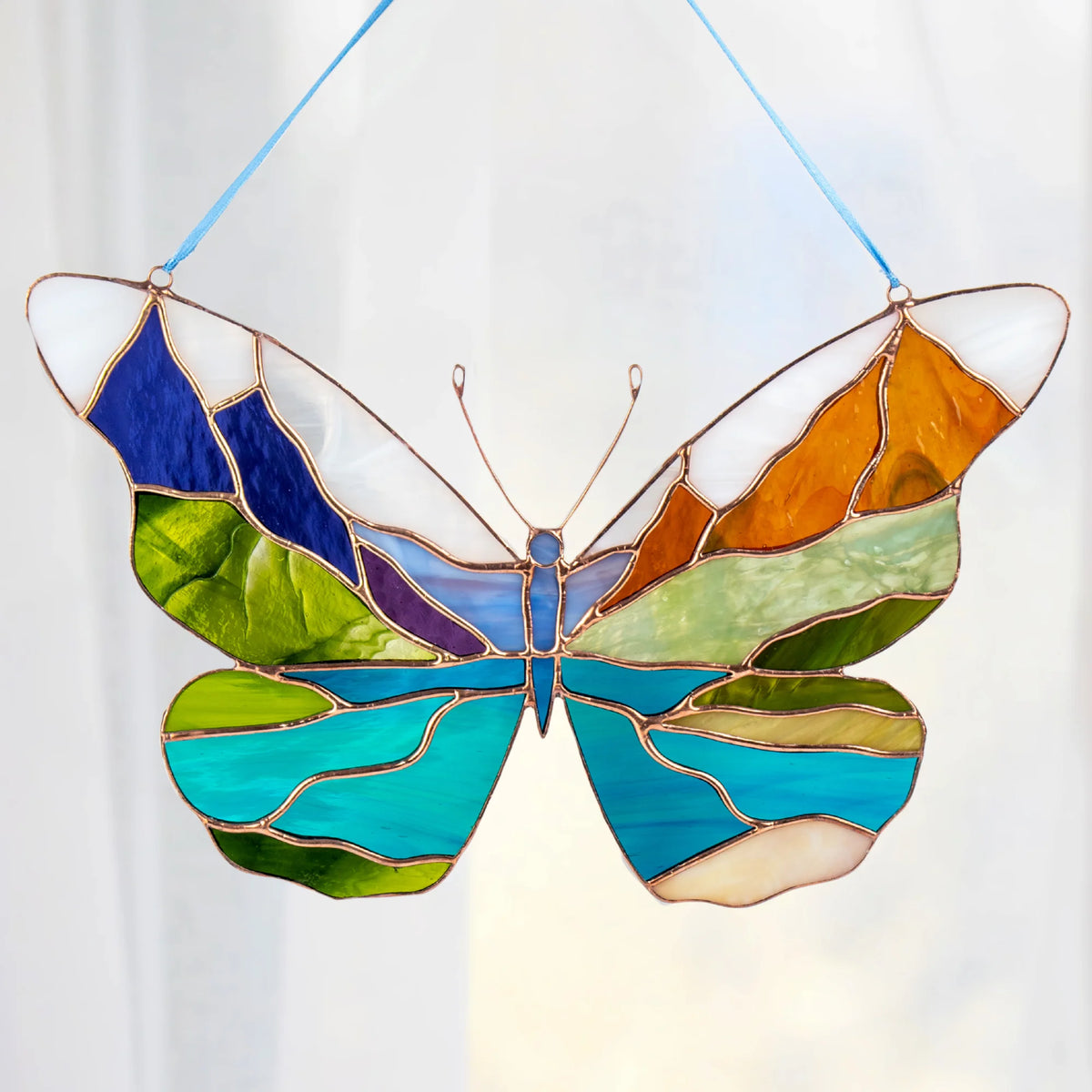 A stained glass butterfly suncatcher hanging from a light blue ribbon. The butterfly features multiple colors of glass including blue, green, orange, purple, and white, separated by copper-colored metal lines.