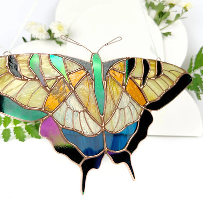 A stained glass butterfly suncatcher, featuring iridescent green, yellow, orange, blue, purple, and black glass panels, hanging against a white background with soft floral props.