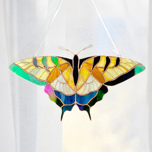 A stained glass swallowtail butterfly suncatcher with vibrant yellow, green, blue, and black sections outlined in copper-colored metal, suspended by thin white strings against a soft white curtain background.