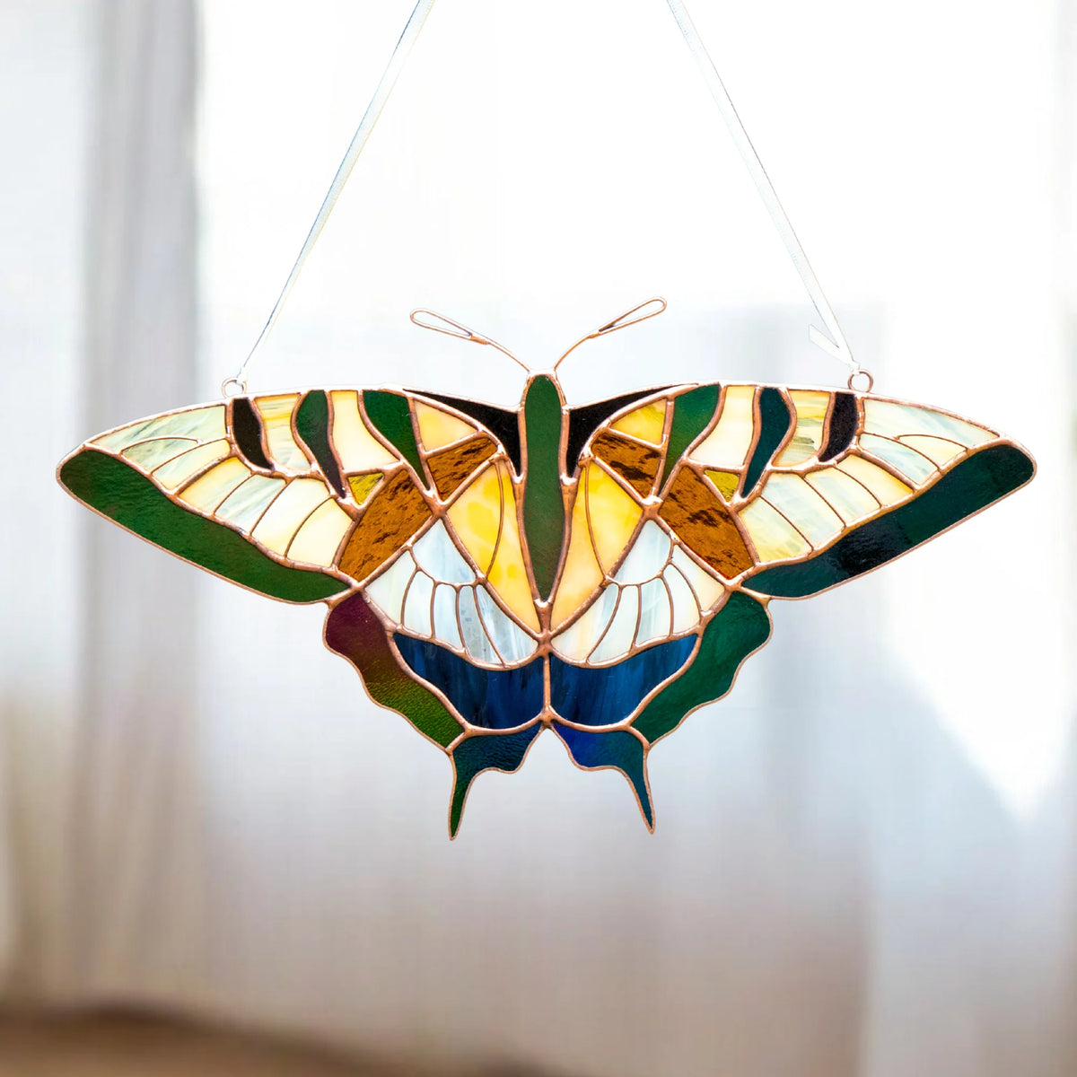 A stained glass butterfly suncatcher with a copper frame, featuring green, yellow, blue, and white sections, hanging against a bright, softly blurred window background.