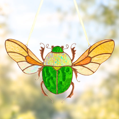 A stained glass insect ornament shaped like a beetle with green body panels, iridescent wings, and brown outlines, suspended by thin white strings against a softly blurred outdoor background.