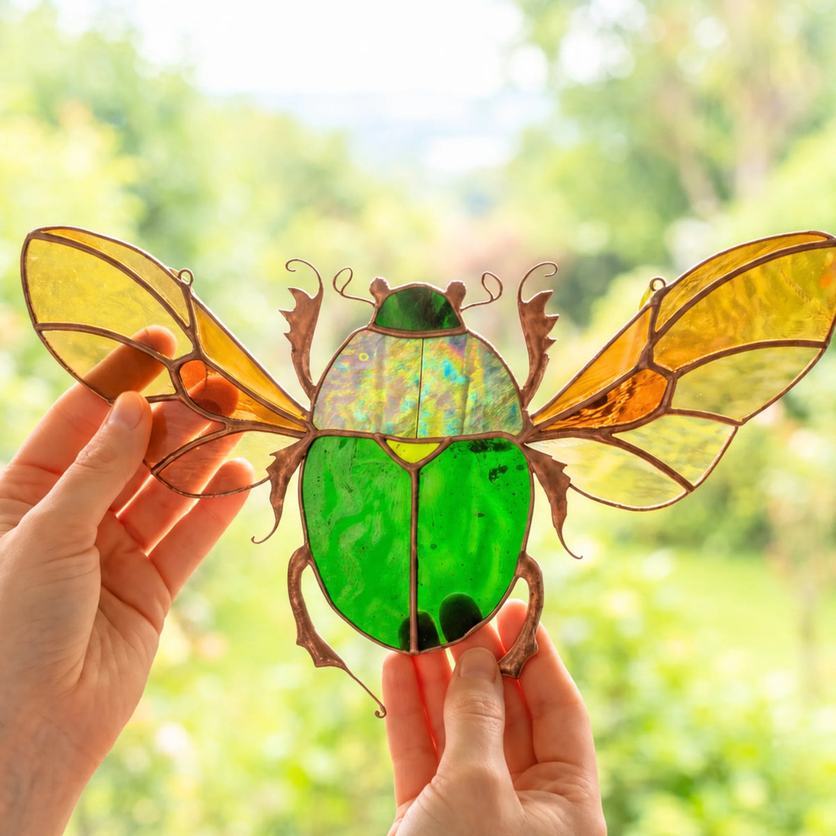 A stained glass scarab beetle ornament with iridescent green and yellow wings, held in two hands against a blurred green garden background.