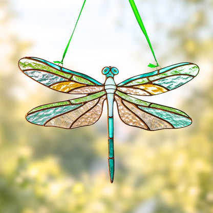 A stained glass dragonfly ornament hanging by a green ribbon, featuring turquoise, green, beige, and amber glass panels with dark metal outlines against a soft, blurred yellow-green background.