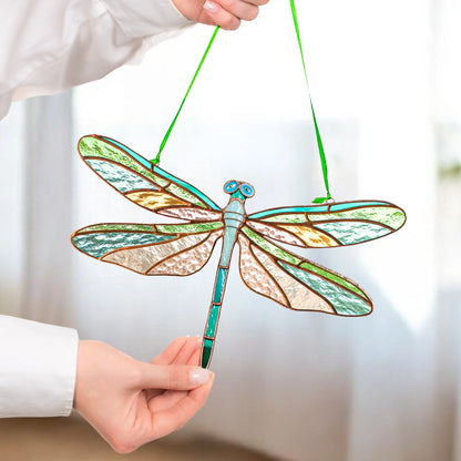 A stained-glass dragonfly ornament with green, blue, and gold-toned details and green hanging strings, held by a person wearing a white long-sleeve shirt against a softly blurred indoor background.
